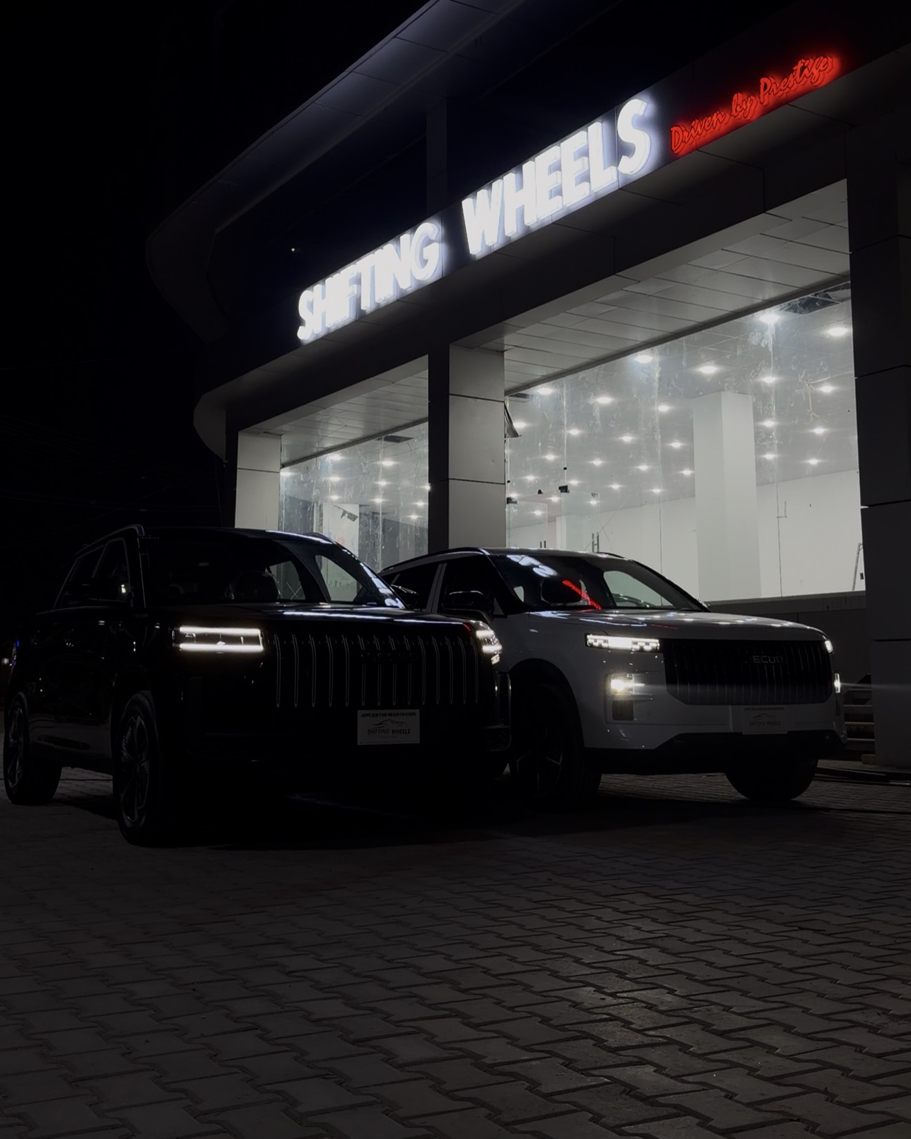 Two SUVs parked in front of the Shifting Wheels showroom at night, under the lit sign